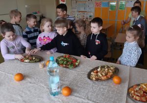 Uczniowie stoją w rzędzie przy stole okrytym białym obrusem. Na stole stoi butelka z wodą oraz leżą cztery pomarańcze i półmiski z koreczkami. Dwoje uczniów sięga do jednego półmiska po koreczki.