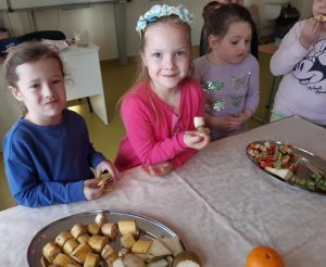 Uczniowie stoją przy stole i jedzą zdrowe przekąski, które nan nim ustawiono.