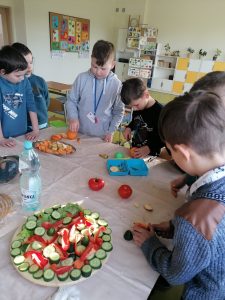 Uczniowie stoją przy dużym stole przykrytym białym obrusem. Dzieci przygotowują zdrowe przekąski. Na stole ustawiony gotowy półmisek z pociętymi ogórkami i papryką, butelka wody i dwa pomidory.