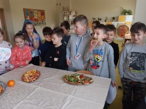 Uczniowie stoją przy dużym stole przykrytym białym obrusem, na którym ustawiono dwa półmiski - jeden z pokrojonymi warzywami a drugi z kawałkami pomarańczy. Jeden z uczniów stojący przy stole je kanapkę.