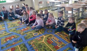 Uczniowie siedzą na kolorowym dywanie i spogladają w jedną stronę.