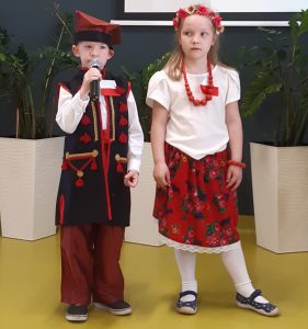 Dwoje uczniów oddziału przedszkolnego w strojach ludowych stoi obok siebie pod jedną ze ścian sali teatralnej. Uczeń trzyma w dłoniach mikrofon.