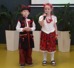 Dwoje uczniów oddziału przedszkolnego w strojach ludowych stoi obok siebie pod jedną ze ścian sali teatralnej. Uczennica trzyma w dłoniach mikrofon. Uczeń stoi wyprostowany.