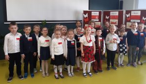 Uczniowie oddziału przedszkolnego w strojach galowych pozują do zdjęcia grupowego podczas apelu na sali teatralnej. Wszystkie dzieci mają przypięte na piersi polskie flagi, a dwoje dzieci stojących po środku ubrana jest w stroje ludowe. W tle widać ścianę sali oraz fragmenty gazetki przgotowanej na temat świąt majowych.