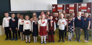 Uczniowie oddziału przedszkolnego w strojach galowych pozują do zdjęcia grupowego podczas apelu na sali teatralnej. Wszystkie dzieci mają przypięte na piersi polskie flagi, a dwoje dzieci stojących po środku ubrana jest w stroje ludowe. W tle widać ścianę sali oraz fragmenty gazetki przgotowanej na temat świąt majowych.