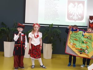 Dwoje uczniów oddziału przedszkolnego w strojach ludowych stoi obok siebie pod jedną ze ścian sali teatralnej. Obok nich dwójka ich kolegów trzyma zieloną mapę Polski z przyklejonymi ważnymi dla kraju ośrodkami. Uczeń trzyma w dłoniach mikrofon. W tle widać tablicę rzutnika na której wyświetla się zdjęcia godła naszego kraju wraz z ważnymi na jego temat informacjami po lewej stronie.
