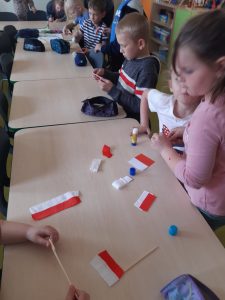 Uczniowie oddziału przedszkolnego podczas tworzenia miniaturowych flag polski. Dzieci siedzą przy wspólnym długim stole, na którym leżą elemnty potrzebne do zbudowania flagi, kleje i piórniki.