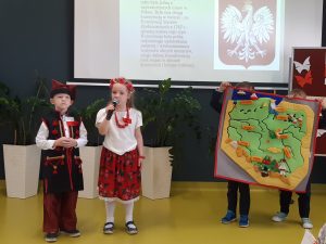 Dwoje uczniów oddziału przedszkolnego w strojach ludowych stoi obok siebie pod jedną ze ścian sali teatralnej. Obok nich dwójka ich kolegów trzyma zieloną mapę Polski z przyklejonymi ważnymi dla kraju ośrodkami. Uczennica trzyma w dłoniach mikrofon. W tle widać tablicę rzutnika na której wyświetla się zdjęcia godła naszego kraju wraz z ważnymi na jego temat informacjami po lewej stronie.