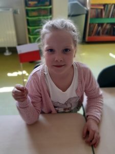 Uczennica oddziału przedszkolnego siedzi przy stoliku i pozuje do zdjęcia trzymając w lewej dłoni zrobioną przez siebie miniaturę polskiej flagi.