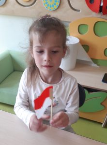 Uczennica oddziału przedszkolnego siedzi przy stoliku i pozuje do zdjęcia trzymając w lewej dłoni zrobioną przez siebie miniaturę polskiej flagi. Dziewczynka ma przymknięte oczy.
