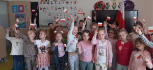 Uczniowie oddziału przedszkolnego pozują do zdjęcia grupowego. W wyciągniętych dłoniach trzymają wykonane przez siebie miniatury polskiej flagi. Dzieci stojące w tyle trzymają w wyciągniętych w górę dłoniach rozpostartą flagę naszego kraju. Dzieci stoją na tle ściany w swojej sali.