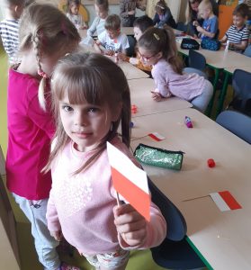 Uczniowie oddziału przedszkolnego podczas tworzenia miniaturowych flag polski. Dzieci siedzą przy wspólnym długim stole ustawionym w literę L, na którym leżą elementy potrzebne do zbudowania flagi, kleje i piórniki. Dziewczynka stojąca na pierwszym planie pozuje do zdjęcia odwrócona tyłem do stolika, trzymając w lewej dłoni gotową flagę.