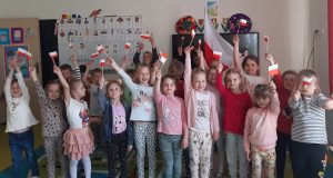 Uczniowie oddziału przedszkolnego pozują do zdjęcia grupowego. W wyciągniętych dłoniach trzymają wykonane przez siebie miniatury polskiej flagi. Dzieci stojące w tyle trzymają w wyciągniętych w górę dłoniach rozpostartą flagę naszego kraju. Dzieci stoją na tle ściany w swojej sali.