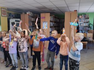 Uczniowie oddziału przedszkolnego pozują do zdjęcia w Bibliotece w Ostaszewie. Dzieci trzymają w wyciągniętych w górę rękach wykonane przez siebie zakładki.