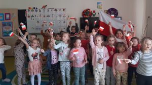 Uczniowie oddziału przedszkolnego pozują do zdjęcia grupowego. W wyciągniętych dłoniach trzymają wykonane przez siebie miniatury polskiej flagi. Dzieci stojące w tyle trzymają w wyciągniętych w górę dłoniach rozpostartą flagę naszego kraju. Dzieci stoją na tle ściany w swojej sali.