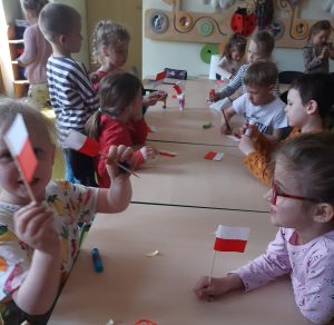 Uczniowie oddziału przedszkolnego podczas tworzenia miniaturowych flag polski. Dzieci siedzą przy wspólnym długim stole, na którym leżą elementy potrzebne do zbudowania flagi. Część uczniów wykonała już swoje zadania i z dumą trzyma w dłoniach gotowe flagi.