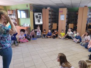 Uczniowie siedzą w półkolu na podlosze Biblioteki w Ostaszewie. Przed nimi stoi Pani Bibliotekarka trzymająca w dłoniach książkę.