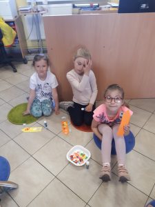 Trzy dziewczynki pozują do zdjęcia siedząc na podłodze Biblioteki w Ostaszewie. Uczennica na pierwszym planie trzyma w dłoniach wykonaną przez siebie zakładkę. Na podłodze obok pozostałych uczennic leżą ich zakładki, kleje oraz pudełeczko z drobnymi elementami, które przyklejały do zakładek.