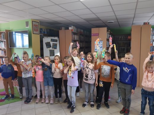 Uczniowie oddziału przedszkolnego pozują do zdjęcia w Bibliotece w Ostaszewie. Dzieci trzymają w wyciągniętych w górę rękach wykonane przez siebie zakładki.