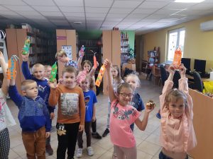 Uczniowie oddziału przedszkolnego pozują do zdjęcia w Bibliotece w Ostaszewie. Dzieci trzymają w wyciągniętych w górę rękach wykonane przez siebie zakładki.
