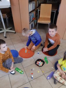 Dzieci siedzą na podłodze Biblioteki w Ostaszewie. Przed nimi leży pudełko zdrobnymi, kolorowymi elementami. Uczniowie wyjmuje te elementy i przyklejają je do wyciętych z papieru książkowych zakładek leżących na podłodze przed nimi.