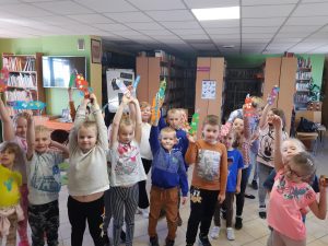 Uczniowie oddziału przedszkolnego pozują do zdjęcia w Bibliotece w Ostaszewie. Dzieci trzymają w wyciągniętych w górę rękach wykonane przez siebie zakładki.
