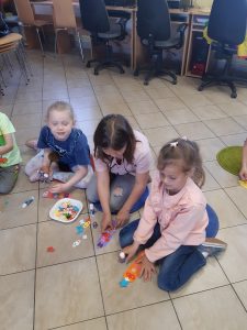 Trzy dziewczynki siedzą obok siebie na podłodze Biblioteki w Ostaszewie. Przed nimi leży pudełko z drobnymi, kolorowymi elementami. Uczennice wyjmują te elementy i przyklejają je do wyciętych z papieru książkowych zakładek leżących na podłodze przed nimi.