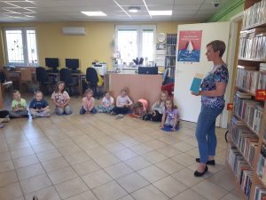 Uczniowie siedzą w półkolu na podlosze Biblioteki w Ostaszewie. Przed nimi stoi Pani Bibliotekarka trzymająca w dłoniach książkę.