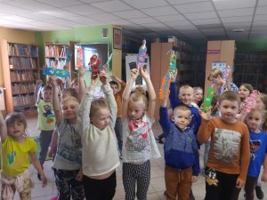 Uczniowie oddziału przedszkolnego pozują do zdjęcia w Bibliotece w Ostaszewie. Dzieci trzymają w wyciągniętych w górę rękach wykonane przez siebie zakładki.