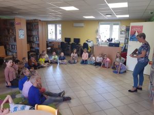 Uczniowie siedzą w półkolu na podlosze Biblioteki w Ostaszewie. Przed nimi stoi Pani Bibliotekarka trzymająca w dłoniach książkę.