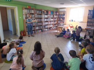 Uczniowie siedzą w półkolu na podlosze Biblioteki w Ostaszewie. Przed nimi stoi Pani Bibliotekarka trzymająca w dłoniach książkę.