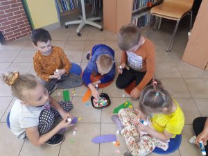 Dzieci siedzą na podłodze Biblioteki w Ostaszewie. Przed nimi leży pudełko z drobnymi, kolorowymi elementami. Uczniowie wyjmują te elementy i przyklejają je do wyciętych z papieru książkowych zakładek leżących na podłodze przed nimi.