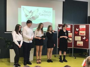 Pięcioro uczniów klasy 8 w strojach galowych stoją w rzędzie pod jedną ze ścian sali teatralnej. Dwie uczennice trzymają w dłoniach mikrofony, a ich koleżanka obok czarną podkładkładkę. W tle widać tablicę rzutnika na której wyświetla się ozdobny napis "Święto Konstytucji" z podobizną orła obok i flagą Polski poniżej. Obok uczniów stoi parawan z gazetką na temat majowych świąt.