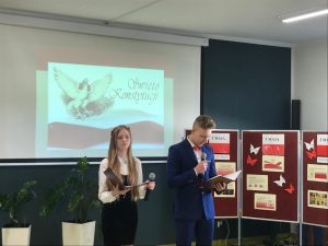 Dwoje uczniów klasy 8 w strojach galowych stoi w rzędzie pod jedną ze ścian sali teatralnej. Uczniowie trzymają w dłoniach mikrofony oraz rozłożone ozdobne teczki. W tle widać tablicę rzutnika na której wyświetla się ozdobny napis "Święto Konstytucji" z podobizną orła obok i flagą Polski poniżej. Obok uczniów stoi parawan z gazetką na temat majowych świąt.