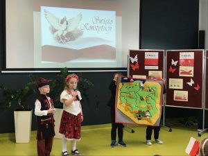 Dwoje uczniów oddziału przedszkolnego w strojach ludowych stoi w rzędzie pod jedną ze ścian sali teatralnej. Obok nich dwójka ich kolegów trzyma zieloną mapę Polski z przyklejonymi ważnymi dla kraju ośrodkami. Uczniowie trzymają w dłoniach mikrofony. W tle widać tablicę rzutnika na której wyświetla się ozdobny napis "Święto Konstytucji" z podobizną orła obok i flagą Polski poniżej. Obok uczniów stoi parawan z gazetką na temat majowych świąt.