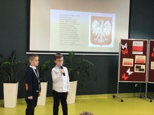 Dwóch uczniów klasy 1 w strojach galowych stoi w rzędzie pod jedną ze ścian sali teatralnej. Jeden z uczniów trzyma w dłoni mikrofon. W tle widać tablicę rzutnika na której wyświetla się polskie godło z tekstem na jego temat zapisanym obok niej. Po lewej stronie uczniów stoi parawan z gazetką na temat majowych świąt.