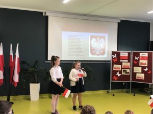 Dwie uczennice klasy 1 w strojach galowych stoi w rzędzie pod jedną ze ścian sali teatralnej. Dziewczyniki trzymają w dłoniach polskie flagi a jedna z nich dodatkowo trzyma w dłoni mikrofon. W tle widać tablicę rzutnika na której wyświetla się polskie godło z tekstem na jego temat zapisanym obok niej. Po lewej stronie uczniów stoi parawan z gazetką na temat majowych świąt.