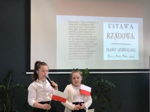 Dwie uczennice klasy 1 w strojach galowych stoją obok siebie pod jedną ze ścian sali teatralnej i trzymają w dłoniach flagi. Jedna z uczennic trzyma w dłoni mikrofon. W tle widać tablicę rzutnika na której wyświetla się pierwsza strona z konstytucji trzeciego maja z napisem: "USTAWA RZĄDOWA. PRAWO UCHWALONE Dnia 3 Maja Roku 1783" z tekstem na jej temat zapisanym obok.