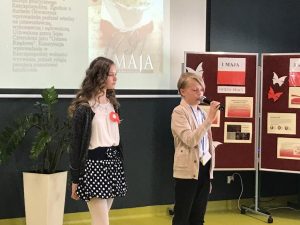 Dwoje uczniów klasy 4 w strojach galowych stoi obok siebie na sali teatralnej Uczeń przemawia przez trzymany w dłoniach mikrofon. W tle widać tablicę rzutnika na której wyświetla się zdjęcie flagi Polski podpisane 3 MAJA oraz tekst dotyczący święta. Po lewej stronie uczniów stoi parawan z gazetką na temat majowych świąt.