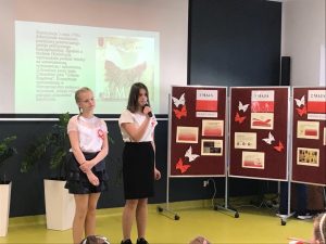 Dwie uczennice klasy 4 w strojach galowych stoją obok siebie na sali teatralnej Jedna z dziewczynek przemawia przez trzymany w dłoniach mikrofon. W tle widać tablicę rzutnika na której wyświetla się zdjęcie polskiej flagi podpisane 3 MAJA oraz tekst dotyczący święta. Po lewej stronie uczennic stoi parawan z gazetką na temat majowych świąt.