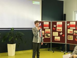 Uczeń w stroju galowym iprzemawia przez trzymany w dłoni mikrofon. W tle biała tablica oraz ustawiony z boku parawan z gazetką zrobioną z okazji majowych świąt.