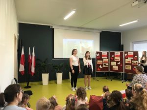 Dwie uczennice stoją na środku sali w galowych strojach. Jedna z nich trzyma mikrofon. W tle biała tablica rzutnika a z boku parawan z gazetką o świętach majowych. Po prawej stronie stoją flagi Polski.