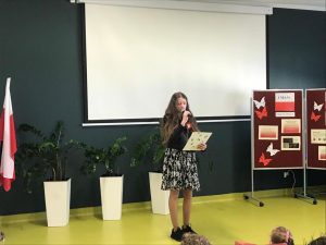 Uczennica w stroju galowym trzyma w jednej dłoni mikrofon a drugiej białą podkładkę. W tle biała tablica oraz ustawiony z boku parawan z gazetką zrobioną z okazji majowych świąt.