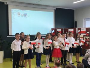 Uczniowie klasy 4 w strojach galowych i przyczepionych biało - czerwonych kotylionach pozują do zdjęcia w sali teatralnej. Dzieci trzymają w dłoniach polskie flagi i stoją na tle białej tablicy rzutnika oraz gazetki przygotowanej z okazji świąt majowych.