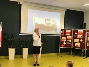 Pani Dyrektor przemawia przez trzymany w dłoni mikrofon. W tle biała tablica na której widoczna jest flaga Polski z godłem powiewająca na wietrze oraz ustawiony z boku parawan z gazetką zrobioną z okazji majowych świąt.