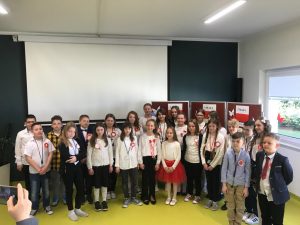 Uczniowie klasy 4 w strojach galowych i przyczepionych biało - czerwonych kotylionach pozują do zdjęcia w sali teatralnej. Stoją na tle białej tablicy rzutnika oraz gazetki przygotowanej z okazji świąt majowych.