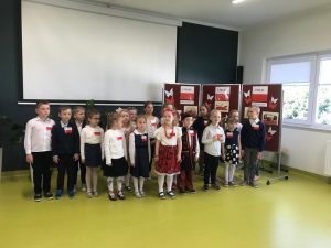 Uczniowie oddziału przedszkolnego w strojach galowych i przyczepionych biało - czerwonych kotylionach pozują do zdjęcia w sali teatralnej. Stoją na tle białej tablicy rzutnika oraz gazetki przygotowanej z okazji świąt majowych.