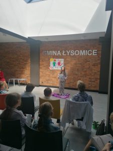 Uczennica w stroju pantery w trakcie recytacji wiersza. Dziewczynka stoi na tle ściany z cegiel z napisem GMINA ŁYSOMICE, pod którym umieszczono grafikę dwojga dzieci z plecakami.