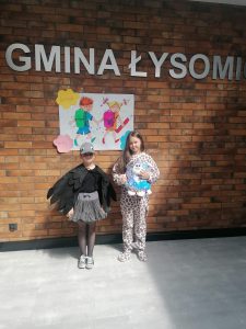 Dwie uczennice pozują do zdjęcia na tle ściany z cegieł znapisem GMINA ŁYSOMICE pod którym umieszczono grafikę dwojga dzieci z plecakami. Dziewczynki ubrane są w swoje stroje sceniczna- jedna z nich ma przyczepione do ramion czarne skrzydła a druga ubrana jest w strój pantery i trzyma w dloniach maskotkę.