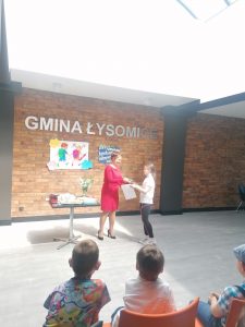 Uczennica odbiera od Pani w czerwonej sukience dyplom. W tle widać ścianę z cegieł a na niej napis GMINA ŁYSOMIC oraz przyczepiona grafika dwojga dzieci z plecakami.